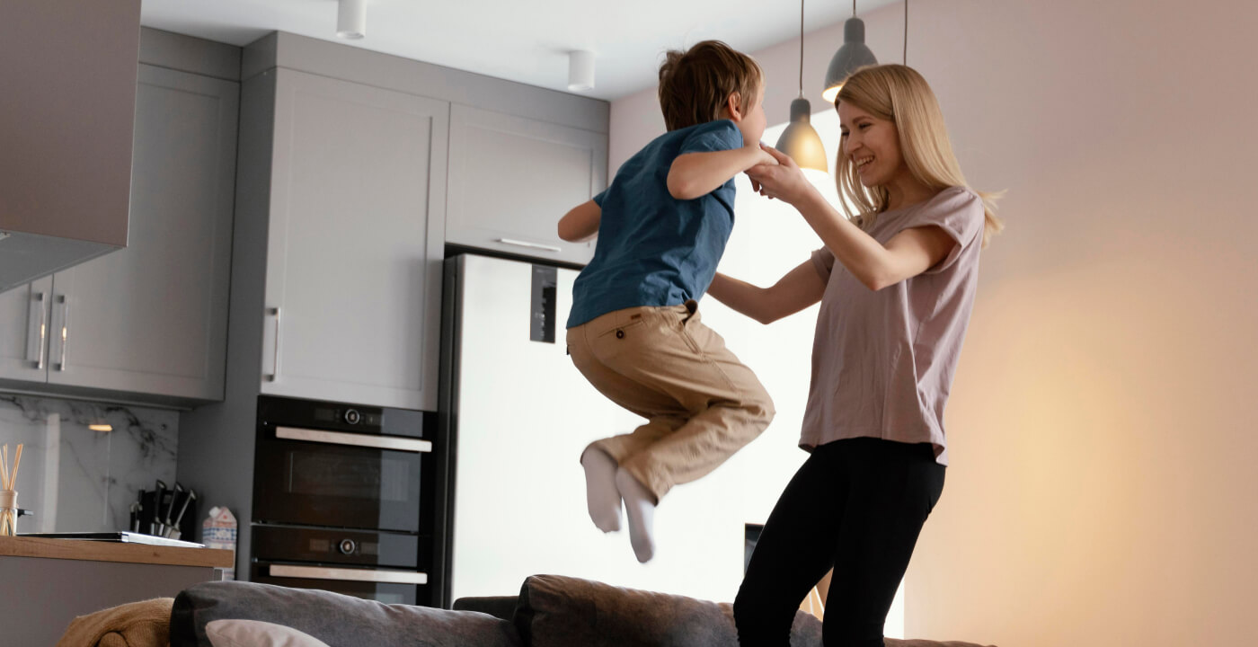 Un bambino piccolo in aria, sollevato giocando dalle mani di un adulto in una cucina moderna, con il bambino che sembra tendere verso una lampada a sospensione.